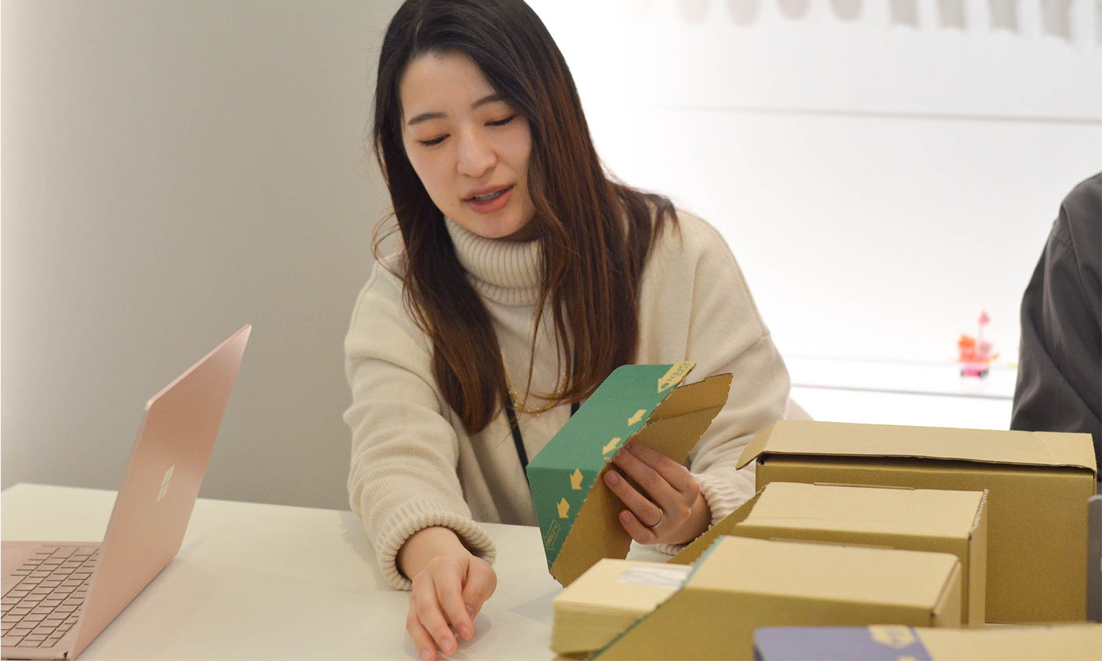 白いセーターを着たカウネット商品開発担当の小原（おはら）さんが左手に商品を手にとり、箱の形状を説明する様子