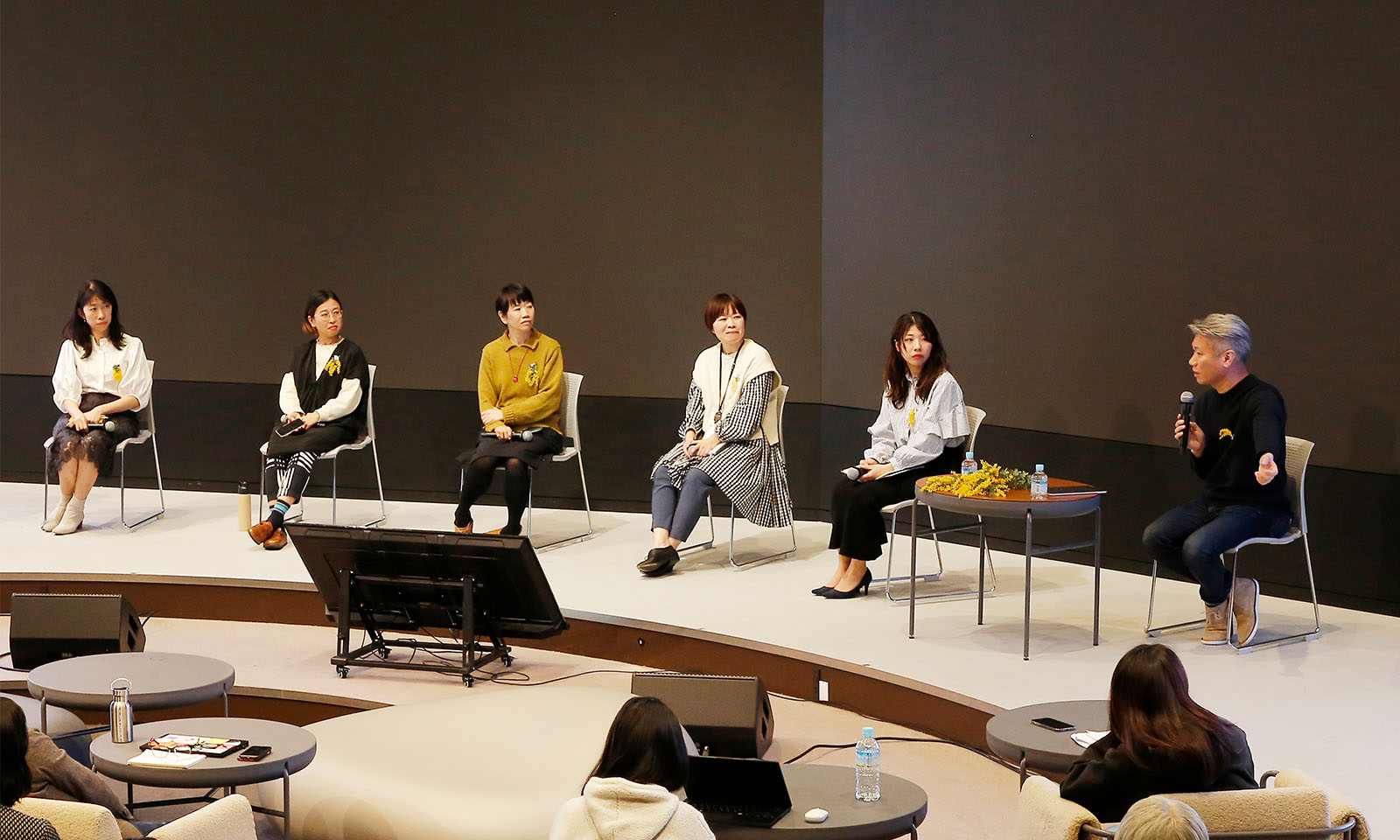 5人のパネリストと越川さんがステージ前方に並んで座り、マイクを持ちながら話す越川さんとその話を真剣に聞くパネリストの様子